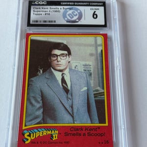 May include: A graded Superman II trading card featuring Clark Kent in a gray suit and glasses. The card is in a protective case and has the text "Clark Kent Smells a Scoop!" and "Topps - #16". The card is graded EX/NM 6.