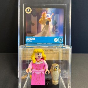 May include: A clear plastic display case with a LEGO minifigure of Aurora from Sleeping Beauty. Aurora is wearing a pink dress with gold stars and has blonde hair. She is holding a small brown owl. The case also includes a card with a picture of Aurora and the text "AURORA Briar Rose 2/5".