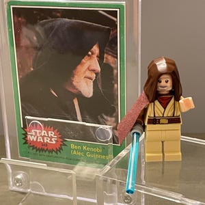 May include: A Star Wars trading card featuring Ben Kenobi (Alec Guinness) in a protective case, with a Lego figure of Obi-Wan Kenobi and a blue lightsaber. The card has a green border and the Star Wars logo.