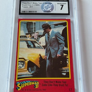 May include: A graded trading card from the Superman II (1980) series, featuring a scene with a yellow taxi cab and a man in a suit. The card is encased in a protective holder and graded as Near Mint 7. The card's text reads "They Don't Make Taxi Cabs Like They Used To!"