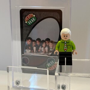May include: A clear acrylic display case holding a Friends-themed trading card with the Central Perk logo. A Lego figure of a character from the show stands next to the card. A small Lego brick structure is in the foreground.