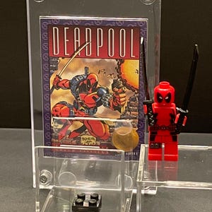 May include: A clear acrylic display case with a red and black Lego Deadpool minifigure. The case also includes a comic book card with the text "DEADPOOL" and a small black Lego brick.