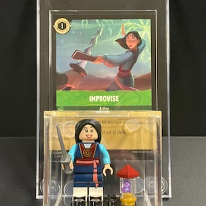 May include: A clear plastic display case with a Lego minifigure of Mulan from the Disney movie Mulan. Mulan is wearing a blue and red outfit and holding a sword. The case also includes a card with the text "IMPROVISE" and a small, clear container with a purple dragon inside.