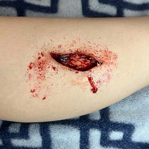 May include: A realistic prosthetic special effects makeup application depicting a deep, open laceration wound on skin. The wound features dark red, textured tissue, bright red blood splatter, and a jagged, irregular opening.