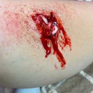 May include: A realistic special effects makeup application depicting a deep, jagged laceration on skin. The wound features textured, dark red simulated blood pooling and dripping, with streaks of opaque white cream filling the center cavity.