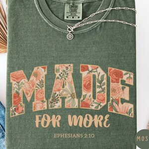 May include: A moss green t-shirt with the words "MADE FOR MORE" in a floral design. The shirt also features the text "Ephesians 2:10". A silver necklace rests on the shirt. The shirt is a Comfort Colors brand.