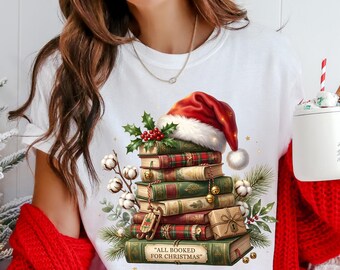 May include: White t-shirt featuring a stack of books with a Santa hat, holly, and cotton accents. The books have green and red covers, and the text "ALL BOOKED FOR CHRISTMAS" is visible.