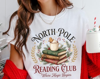 May include: White t-shirt with a festive design featuring the words "North Pole Reading Club" and "Where Magic Begins." The graphic includes a stack of books, a mug of hot cocoa with a gingerbread man, and a wreath. The shirt is a crew neck.