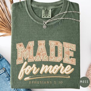 May include: A moss green t-shirt with the words "MADE for more" in a floral design. The text "Ephesians 2:10" is below the main text. A silver necklace with a pendant is draped across the shirt.