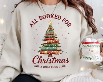 May include: Cream-colored sweatshirt with a Christmas tree design made of books. The text reads "All Booked For Christmas" and "Holly Jolly Book Club" in red. A gold star tops the tree.