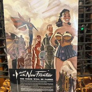 Puede incluir: Una portada de cómic con superhéroes de DC Comics, incluidos Superman, Wonder Woman y The Flash. La portada tiene un estilo vintage con el título "Absolute DC The New Frontier" y el texto "The Torch Will Be Passed".