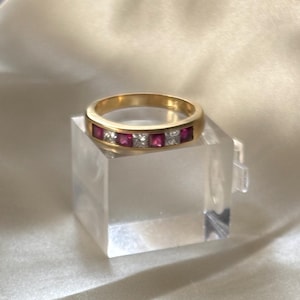 May include: A gold band ring featuring an alternating channel-set row of square-cut red rubies and clear white diamonds. The ring is polished and has a smooth, rounded exterior surface, showcasing a series of three red stones and two white stones.