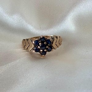 Puede incluir: Un anillo dorado con un grupo de pequeñas piedras preciosas de color azul oscuro dispuestas en forma de flor. La banda del anillo tiene un diseño de chevrón a cada lado del grupo de piedras preciosas. El anillo está sobre un fondo blanco y sedoso.