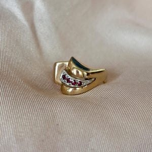 Peut inclure: Une bague en or au design unique. La bague présente une section incurvée avec de petites pierres précieuses rouges et un accent de couleur argentée. La bague a une finition polie et est posée sur un fond neutre.