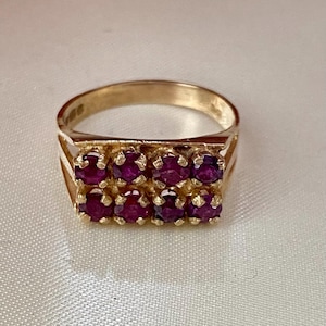 May include: A gold ring with a rectangular setting, holding two rows of deep red gemstones. The ring's design is classic, with a polished finish that highlights the gemstones' colour and sparkle. A timeless piece of jewellery, suitable for any occasion.