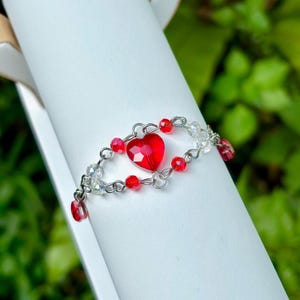 May include: A handmade bracelet featuring a faceted red heart pendant at the center, surrounded by red and clear crystal beads. The bracelet is strung on a silver-colored wire, creating a delicate and romantic design.