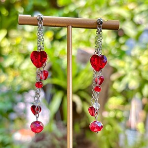 May include: Dangling earrings with red heart-shaped and round beads. The earrings are made of silver-colored chains and are displayed on a gold-colored stand. The background is a blurred view of green foliage.