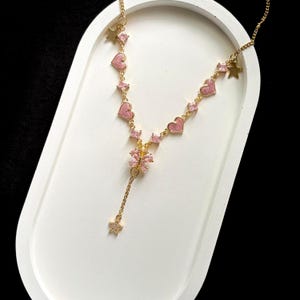 Puede incluir: Un collar de oro con un colgante de mariposa rosa y dijes de corazón y estrella rosa.