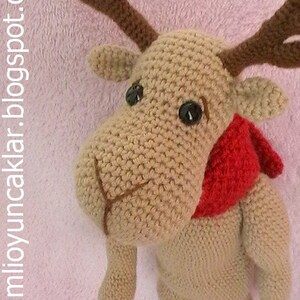 May include: A crocheted reindeer toy with brown antlers, brown eyes, and a red scarf. The reindeer is standing on a pink fuzzy surface.