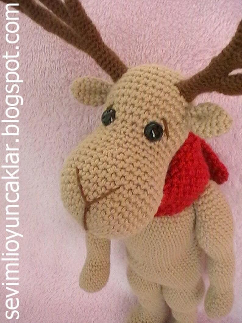 May include: A crocheted reindeer toy with brown antlers, brown eyes, and a red scarf. The reindeer is standing on a pink fuzzy surface.