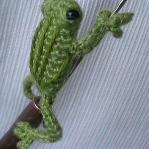 May include: A green crocheted frog with black eyes, sitting on a brown wooden stick. The frog is holding a silver crochet hook in its right hand.