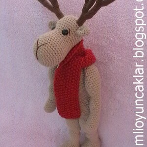 May include: A crocheted reindeer toy with brown antlers and a red scarf. The reindeer is standing on a pink fuzzy surface.