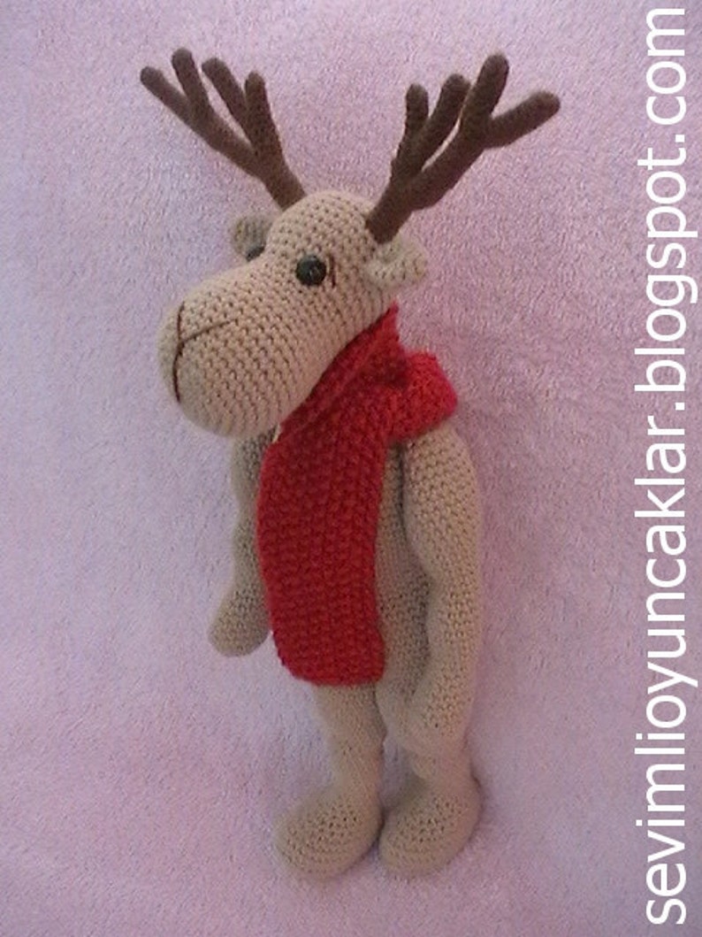May include: A crocheted reindeer toy with brown antlers and a red scarf. The reindeer is standing on a pink fuzzy surface.