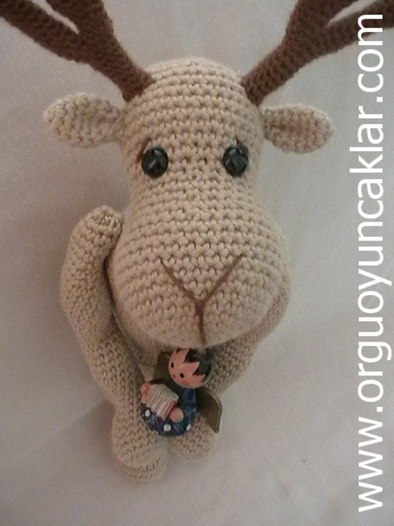 May include: A crocheted reindeer with brown antlers holding a small wooden figurine of a person playing an accordion. The reindeer is beige and has black eyes.