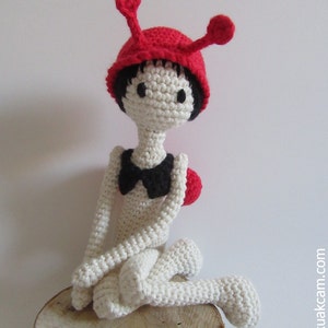 May include: A crocheted doll with white yarn and a red hat with antennas. The doll is wearing a black bow and is sitting on a wooden surface.