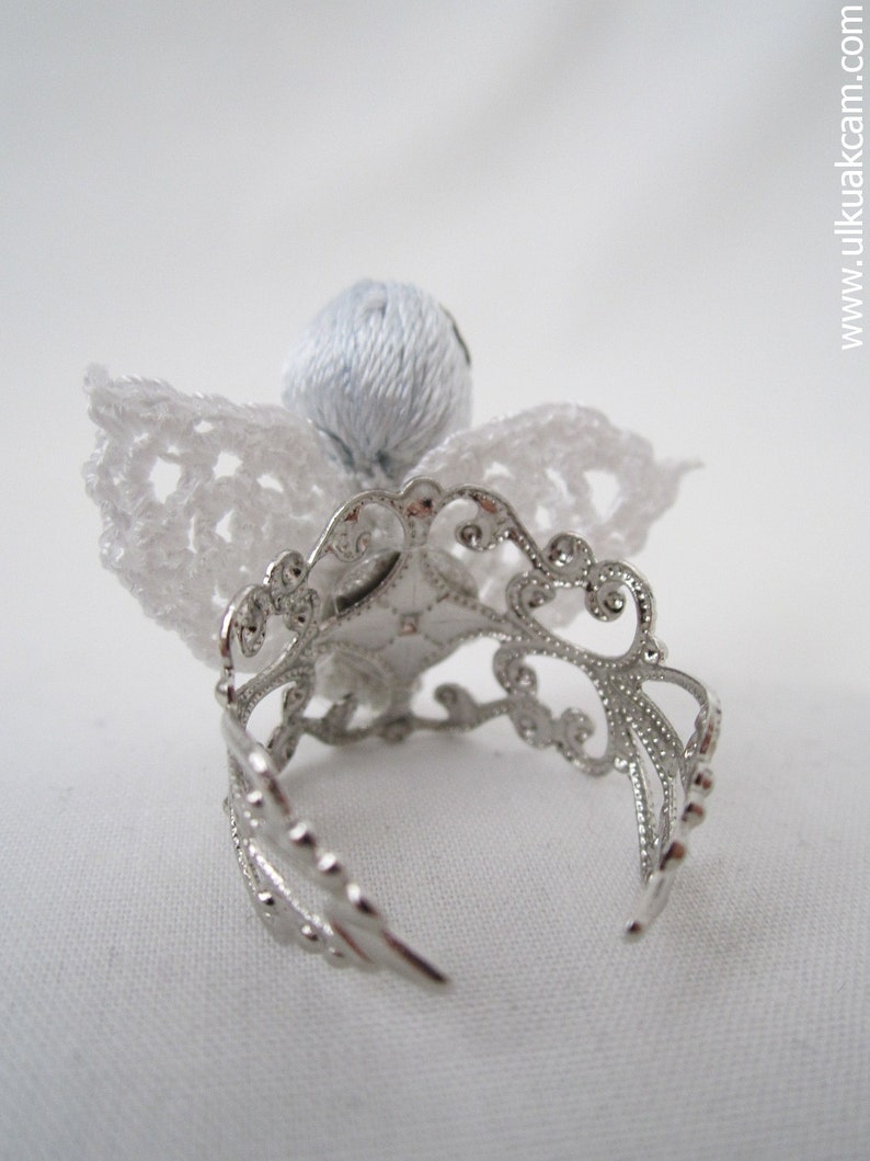 Puede incluir: Un &aacute;ngel de crochet blanco con un anillo de plata. El &aacute;ngel tiene una cabeza y alas de crochet blanco.