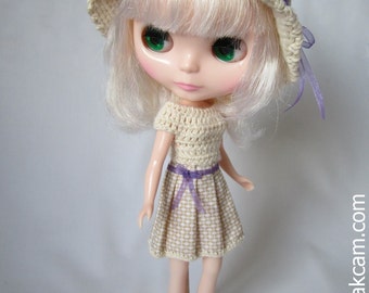 May include: A doll with blonde hair wearing a crocheted dress and hat with a purple ribbon. The doll is wearing pink shoes and is standing on a white platform.