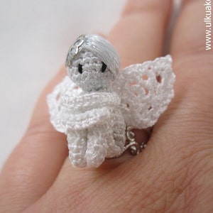 Puede incluir: Un anillo de &aacute;ngel de crochet blanco con una banda de anillo de plata. El &aacute;ngel lleva un vestido y alas de crochet blanco.