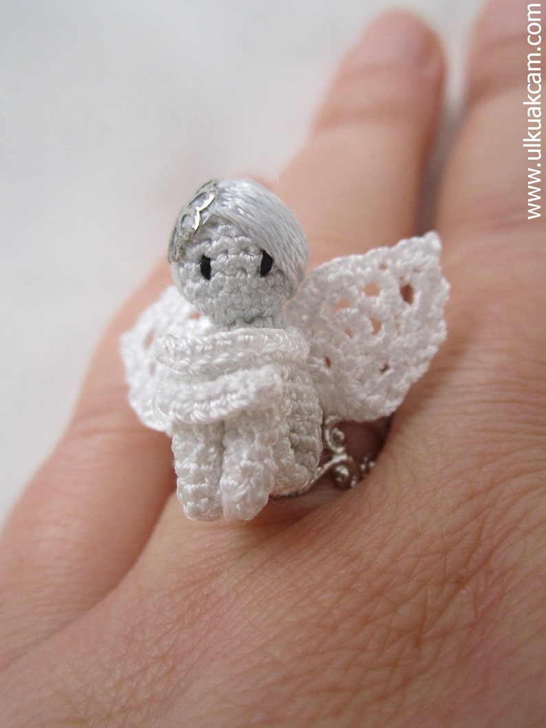Puede incluir: Un anillo de &aacute;ngel de crochet blanco con una banda de anillo de plata. El &aacute;ngel lleva un vestido y alas de crochet blanco.