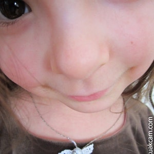 Puede incluir: Una ni&ntilde;a con cabello casta&ntilde;o y ojos marrones lleva una camisa marr&oacute;n y un collar de plata con un colgante de &aacute;ngel de crochet blanco.