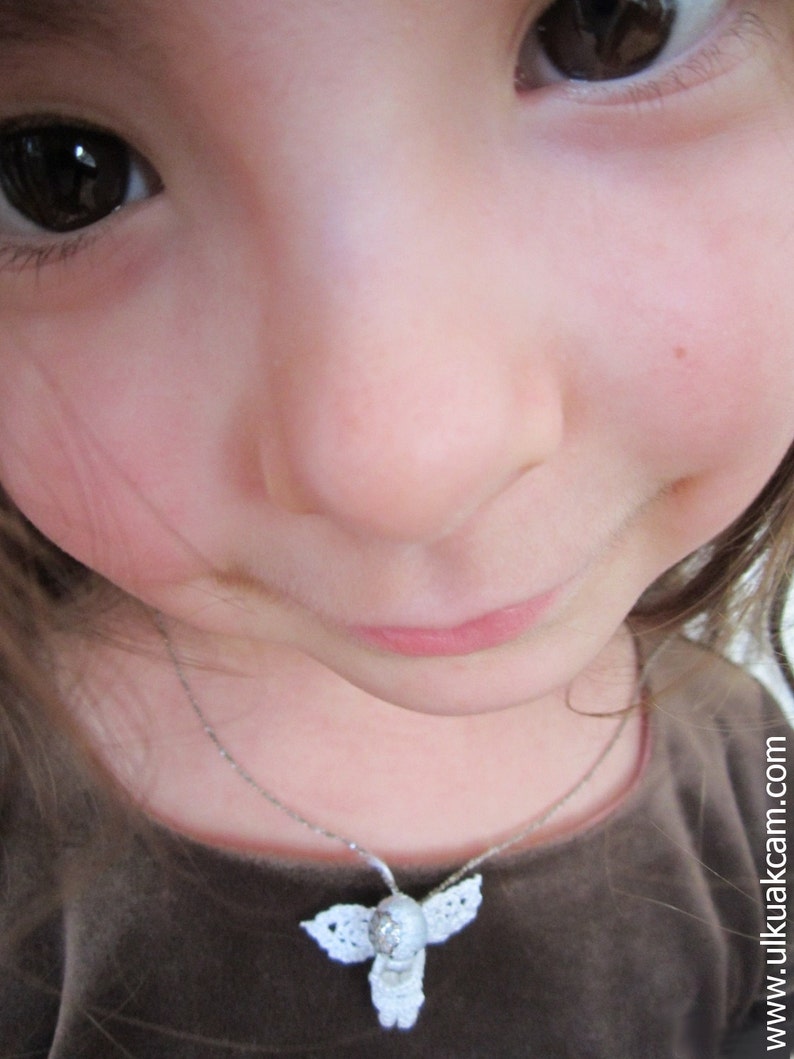 Puede incluir: Una ni&ntilde;a con cabello casta&ntilde;o y ojos marrones lleva una camisa marr&oacute;n y un collar de plata con un colgante de &aacute;ngel de crochet blanco.