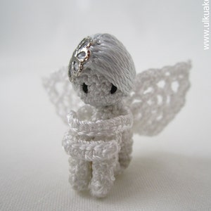 Puede incluir: Una peque&ntilde;a figura de &aacute;ngel de crochet blanco con un halo y alas plateadas. El &aacute;ngel tiene las manos juntas frente a su pecho.