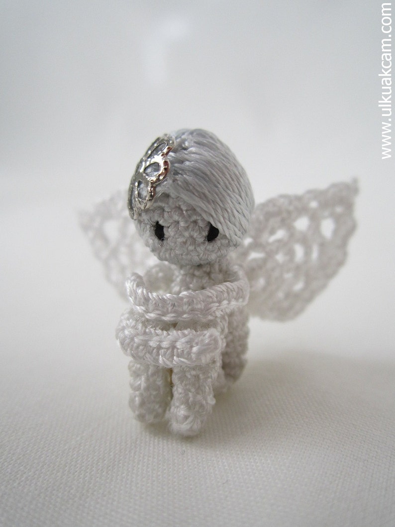 Puede incluir: Una peque&ntilde;a figura de &aacute;ngel de crochet blanco con un halo y alas plateadas. El &aacute;ngel tiene las manos juntas frente a su pecho.