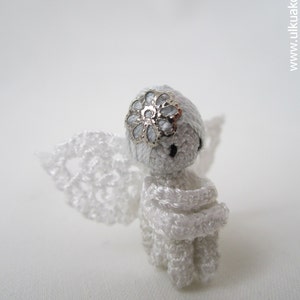 Puede incluir: Una figurita de &aacute;ngel de crochet blanco con un acento de flor plateada en la cabeza. El &aacute;ngel tiene un vestido y alas de crochet blanco.