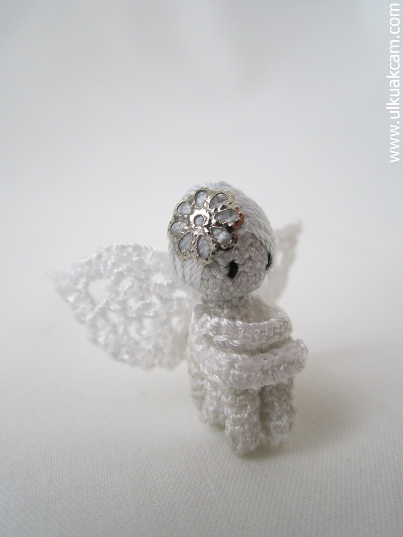 Puede incluir: Una figurita de &aacute;ngel de crochet blanco con un acento de flor plateada en la cabeza. El &aacute;ngel tiene un vestido y alas de crochet blanco.