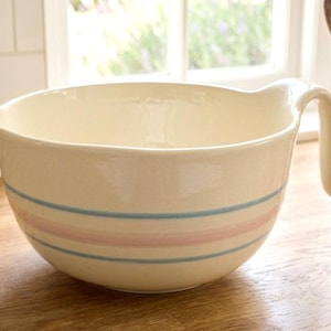 Può includere: Ciotola per mescolare in ceramica color crema con manico e beccuccio. La ciotola presenta due strisce orizzontali azzurre e una rosa. È appoggiata su una superficie di legno.