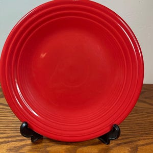 May include: A vibrant red ceramic plate with concentric rings near the rim. The plate is displayed on a black stand, showcasing its bright color and simple design. This plate is suitable for serving food.