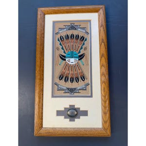 Peut inclure: Une peinture sur sable rectangulaire dans un cadre en bois de chêne clair présentant un motif central de masque turquoise et blanc entouré de plumes brunes et noires. Sous l'œuvre, un petit concho ovale en métal argenté est centré sur un passe-partout.