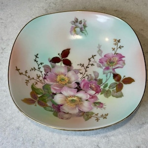 May include: Square porcelain dish with a gold rim, featuring a floral design of pink roses, green leaves, and brown accents. The background has a soft gradient of pink and green hues.