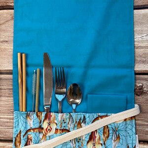 May include: A turquoise fabric cutlery roll with a white strap and a colorful pattern of feathers and dreamcatchers. The roll contains a fork, knife, spoon, two bamboo straws, and a cleaning brush.