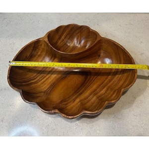 Puede incluir: Un cuenco de madera para servir con tres compartimentos y un borde festoneado. El cuenco es de color marrón cálido y mide aproximadamente 43 cm de largo. El cuenco está vacío.