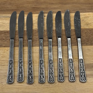 Puede incluir: Un juego de ocho cuchillos de mantequilla vintage. Los cuchillos tienen un acabado plateado con un patrón decorativo en los mangos. Las hojas son curvas y los cuchillos están colocados sobre una superficie de madera.