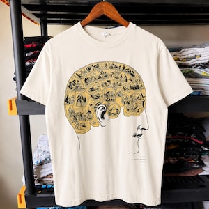 May include: A cream-colored short-sleeve t-shirt featuring a black and yellow phrenology-inspired graphic of a human head profile. The brain area is divided into labeled sections with small illustrations and the text "THERE'S A WHOLE WORLD IN HERE."