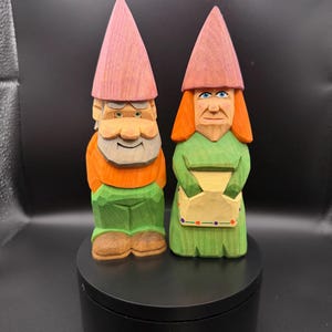 May include: Two hand-carved wooden garden gnomes. The gnome on the left has an orange shirt, green pants, and a pink hat. The gnome on the right has a green dress, orange hair, and a pink hat. Both gnomes have detailed facial features.