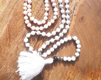 May include: Two beaded necklaces with a tassel. One necklace features peach-colored beads, while the other has white and gray beads. The necklaces are adorned with metal charms, including a Hamsa hand and a spiral design.