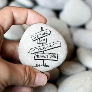 Könnte beinhalten: Ein glatter, hellfarbener Stein mit schwarzer Tinte. Der Text lautet "It's time for a new adventure" in einem Wegweiser-Design. Der Stein wird in einer Hand gehalten, mit anderen Steinen im Hintergrund.